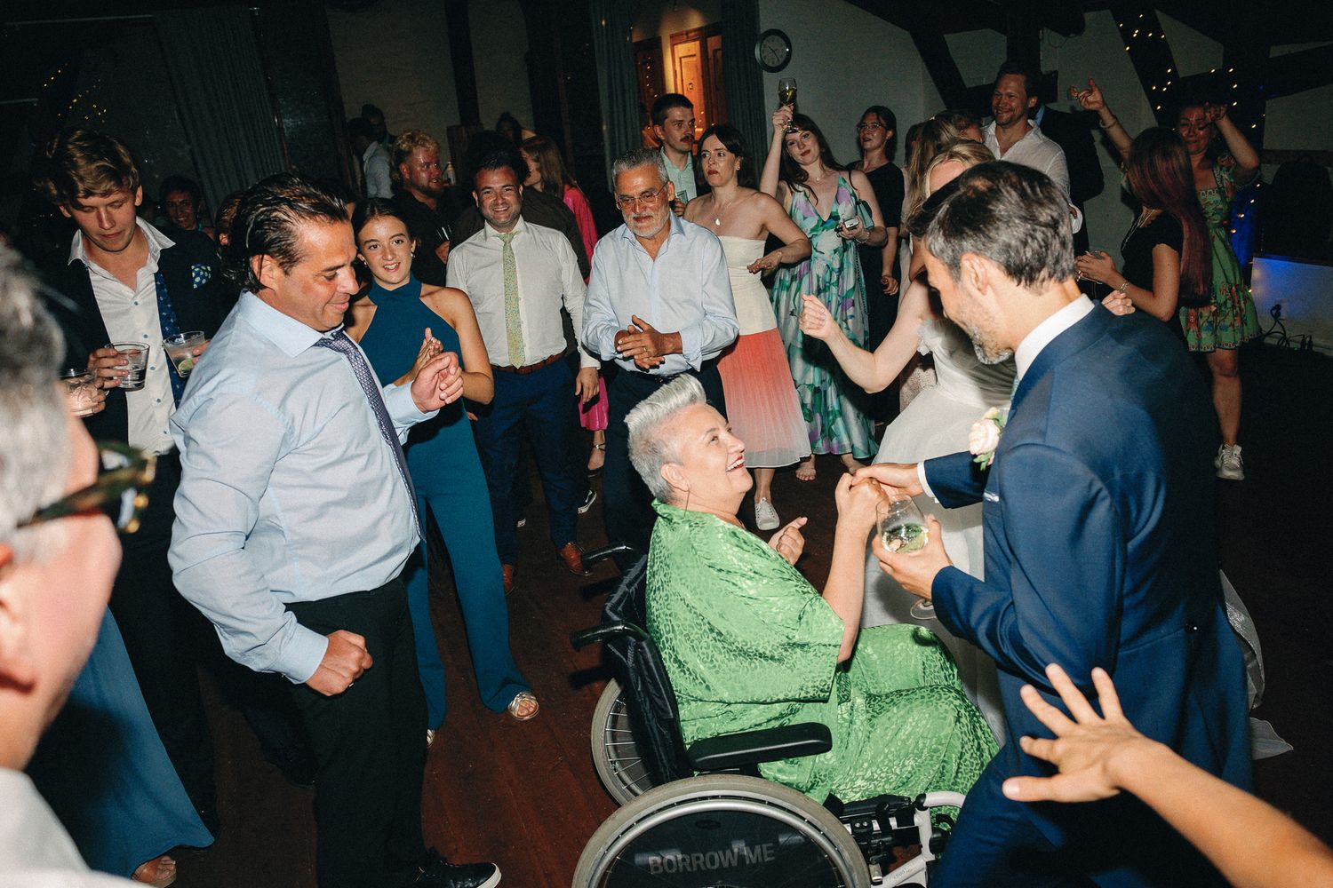 Guests dancing joyfully at a wedding reception, including an elderly person in a wheelchair joining the celebration.