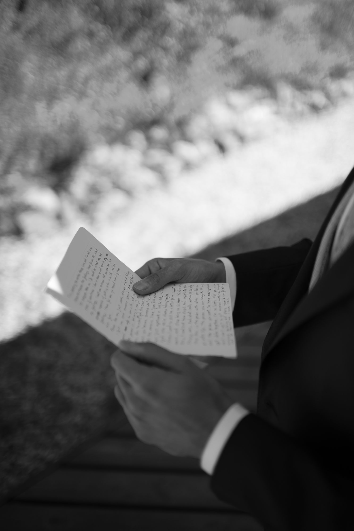 Black and white photo of a groom in a suit reading handwritten wedding vows from a folded paper outdoors.