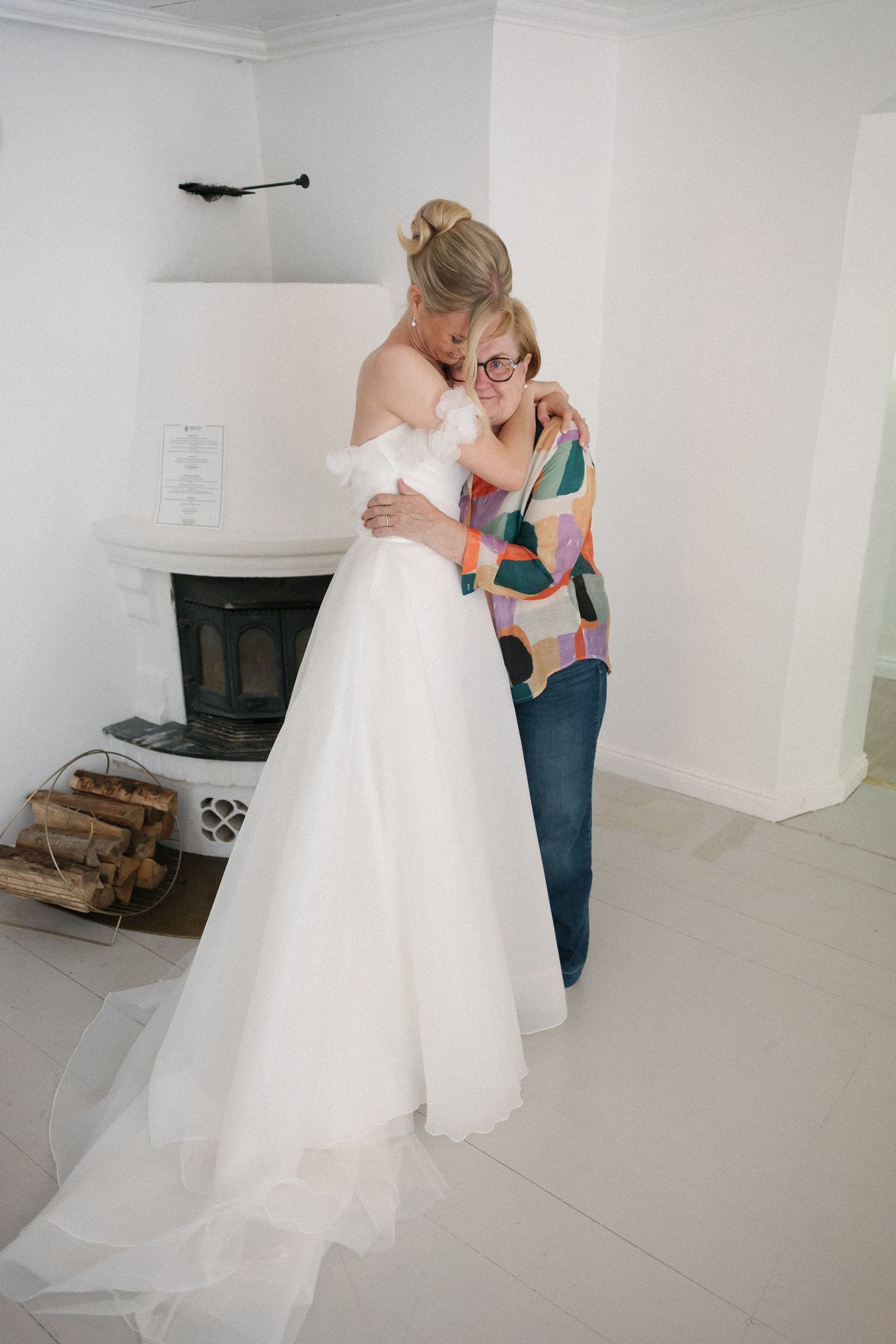 Bride in white off-shoulder gown embracing a woman in colorful shirt near fireplace during wedding preparation.