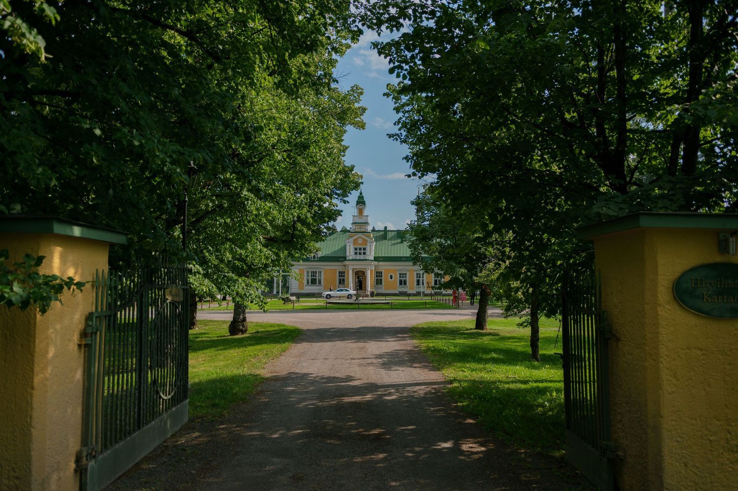 Tree-lined pathway leading to a white building with columns in a lush garden setting.