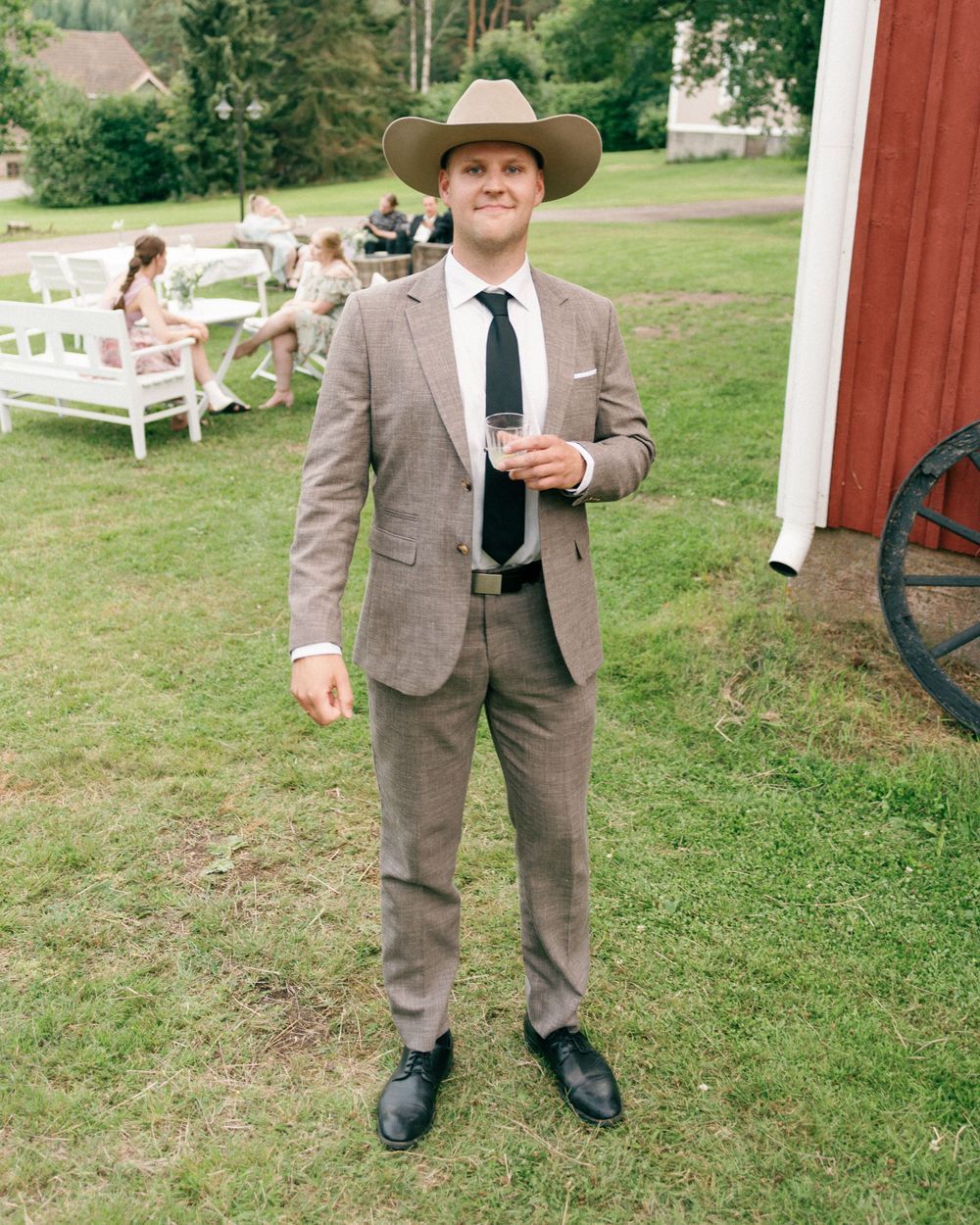 Person in a brown suit and cowboy hat stands on grass near a red barn.