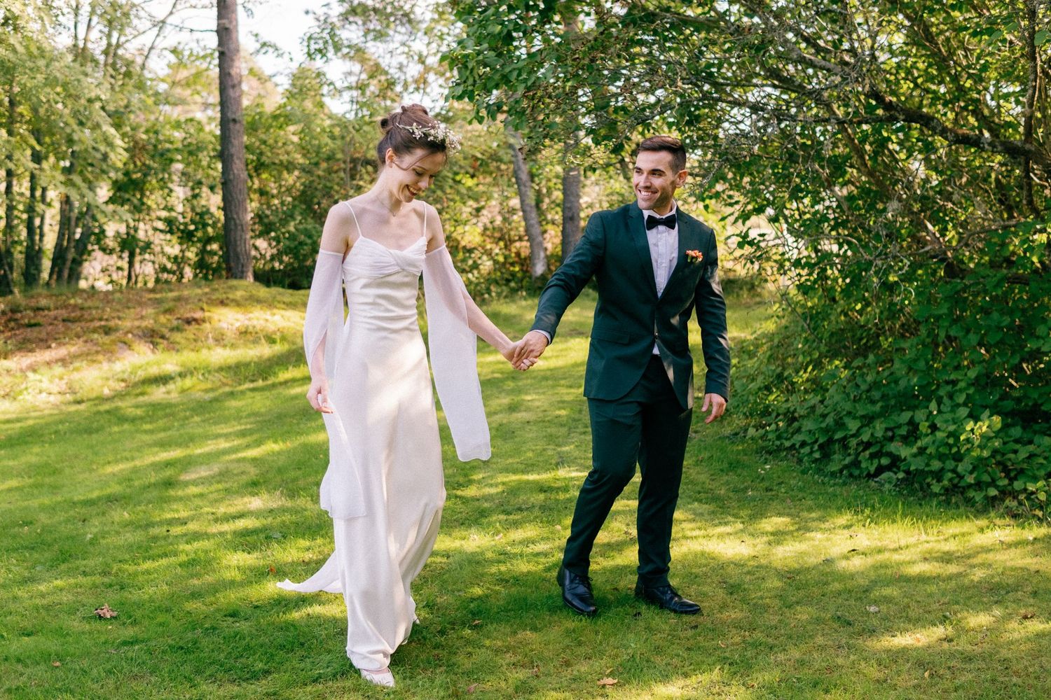 Playful wedding couple dancing and twirling on a sunny lawn surrounded by trees.