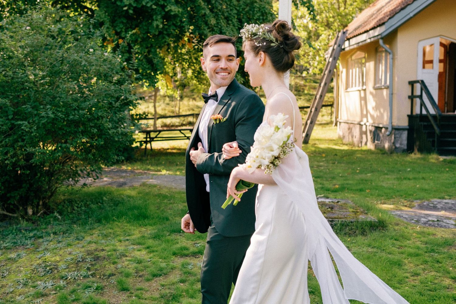 An outdoor wedding ceremony takes place on a lush green lawn with a historic yellow building in the background.