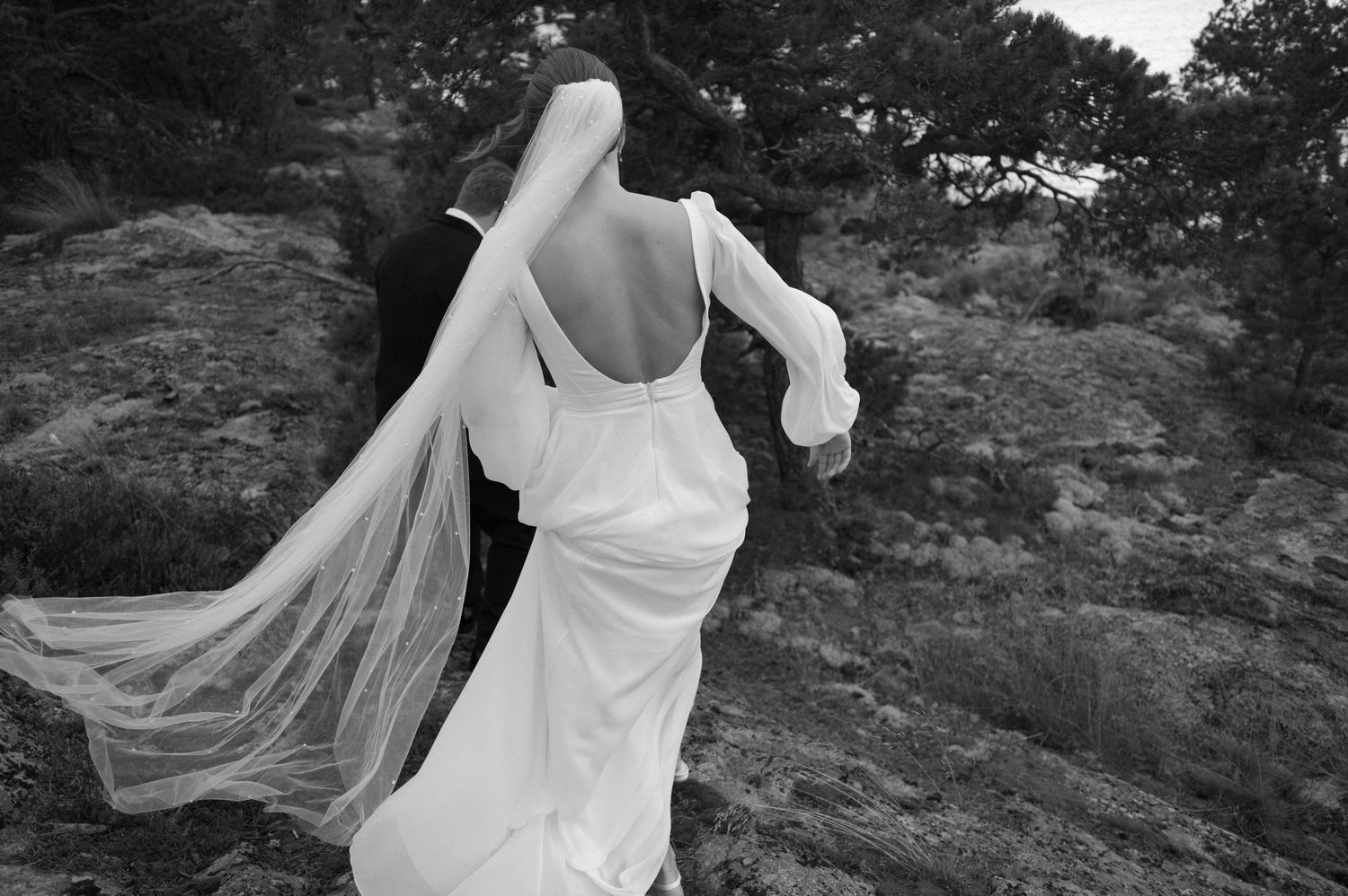 A bride in a flowing white wedding dress walks along a scenic mountain path with her veil trailing in the wind.