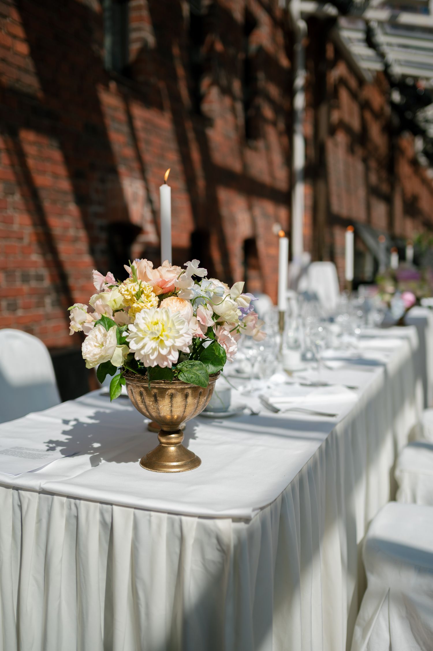 Elegant outdoor wedding table with white linens, gold floral centerpiece, candle, and glassware against brick wall.