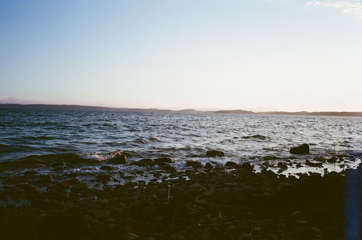 Rocky shoreline with choppy ocean waves at sunset, distant mountains on horizon under clear sky.