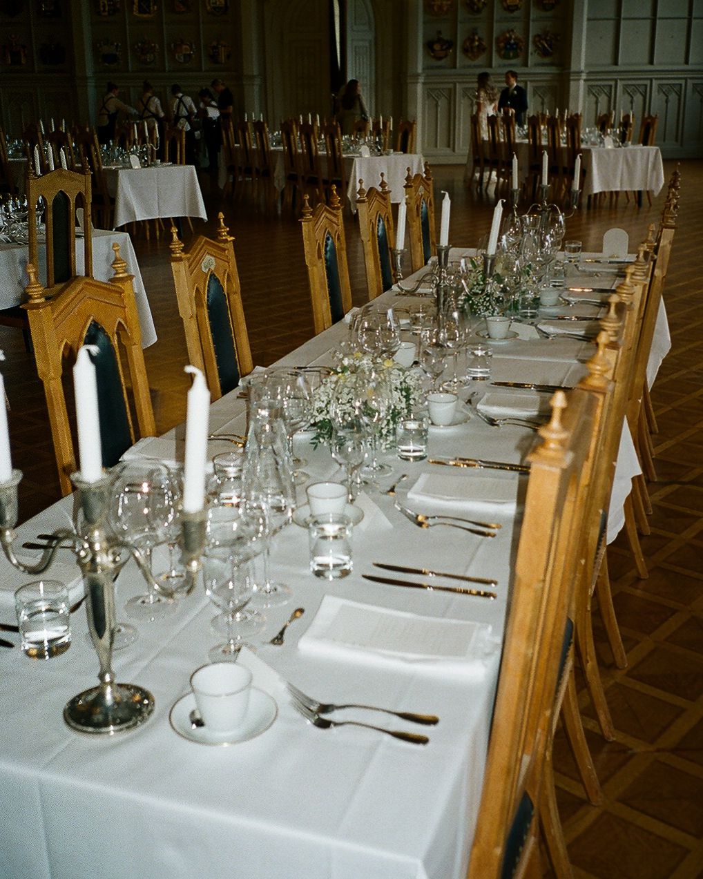 Elegant long dining table set with white linens, candles, glassware, and floral centerpieces in a grand hall.