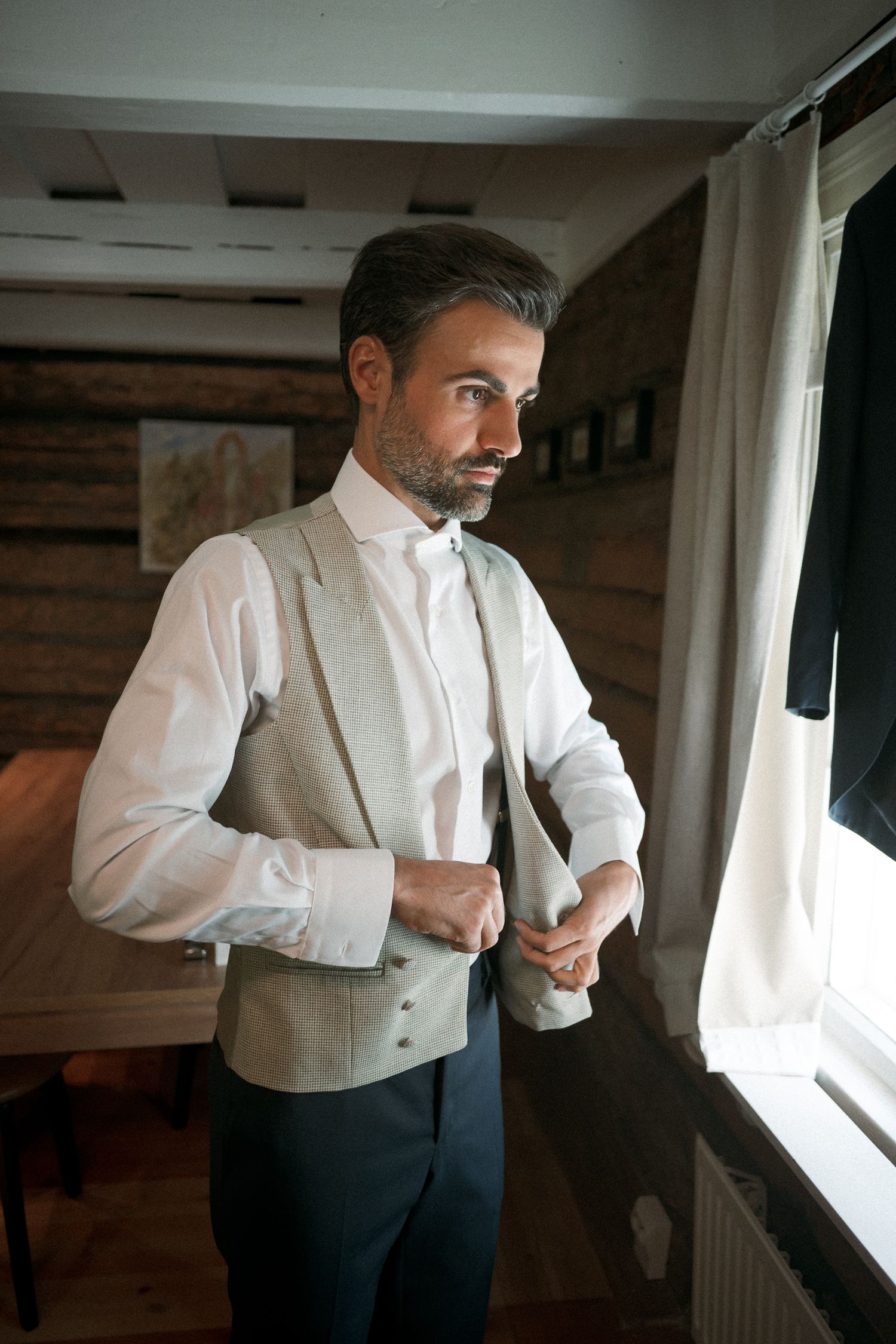 A well-dressed man in a white shirt and gray vest buttoning up near a window in a rustic wooden room.