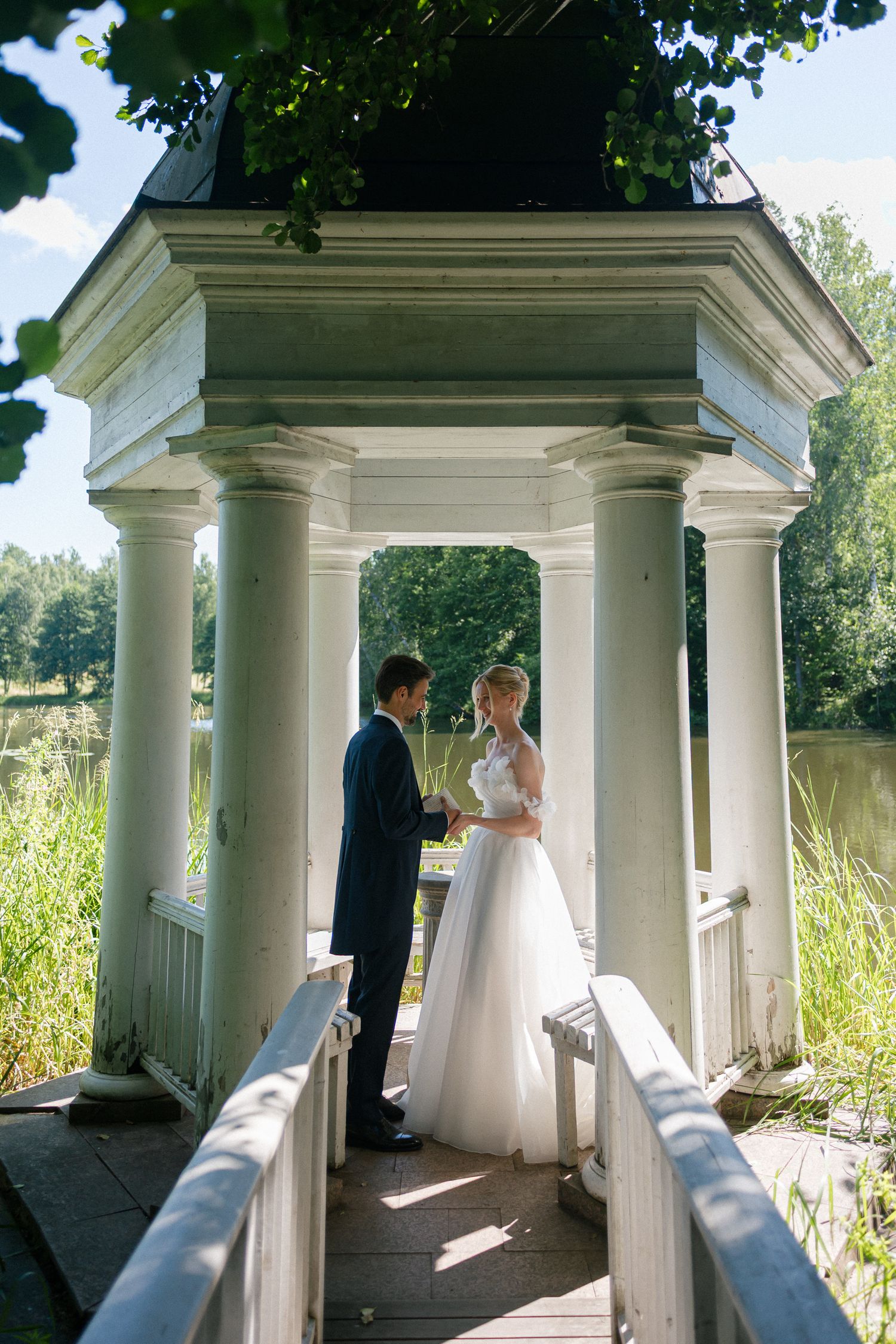 Bride and groom holding hands in a white gazebo by a lake, surrounded by lush greenery on a sunny wedding day.