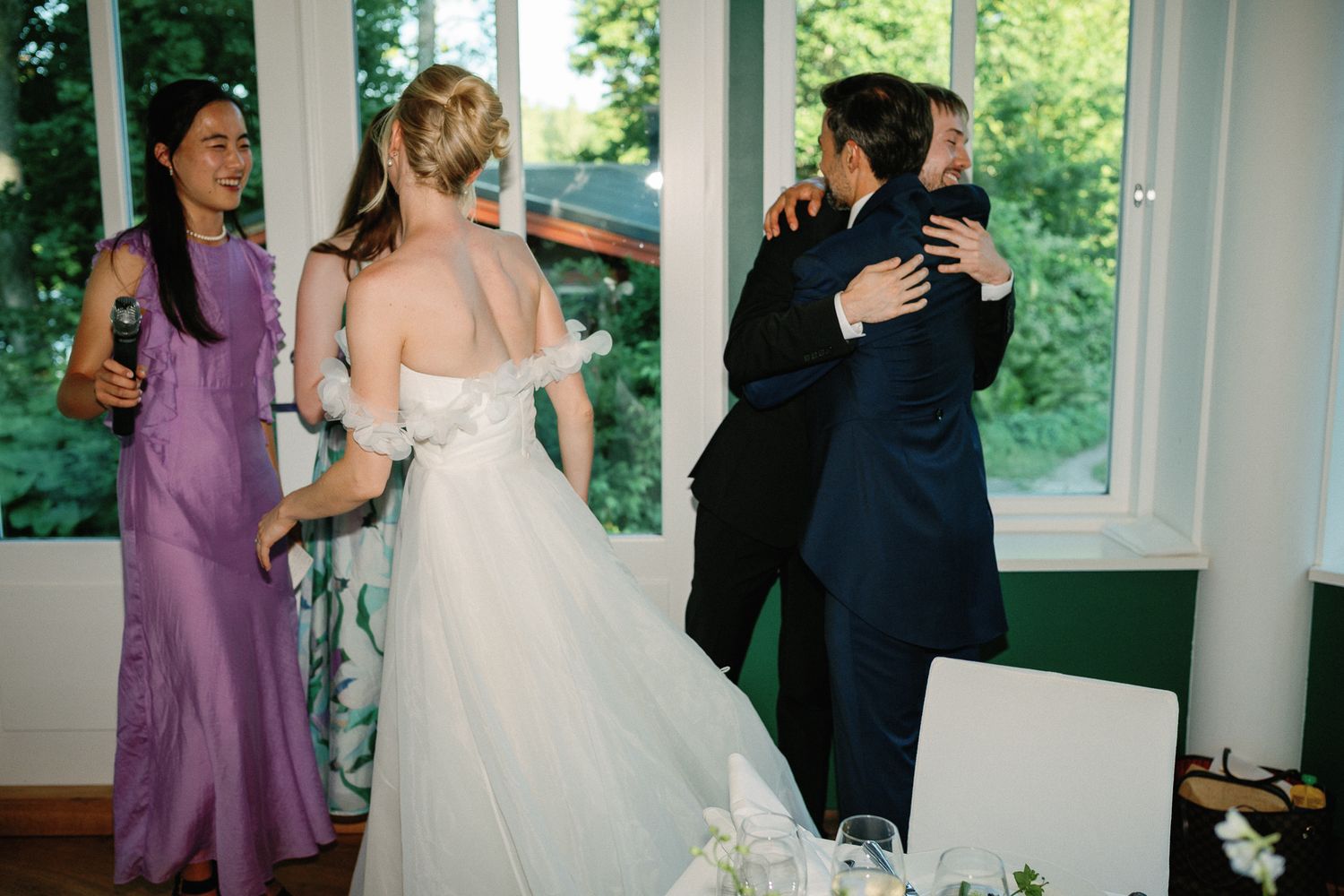 Bride in white gown and guests in purple and navy attire celebrate indoors near large windows at a wedding reception.