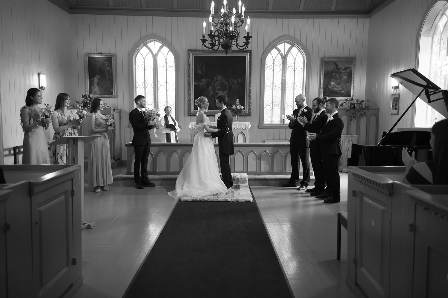 Black and white photo of a wedding ceremony inside a classic church with chandelier, bridal party, and altar.