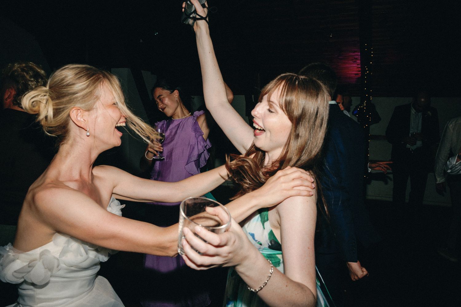 Two women dancing joyfully at a wedding reception, one in a white bridal gown, arms raised in celebration.