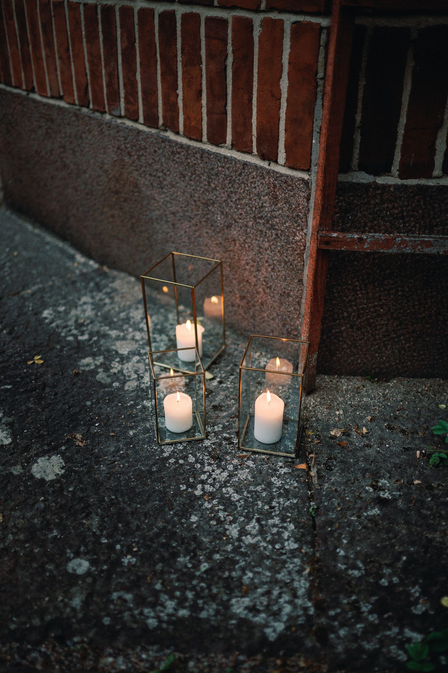 Two gold geometric glass candle holders with lit white pillar candles placed on stone steps near a brick wall.