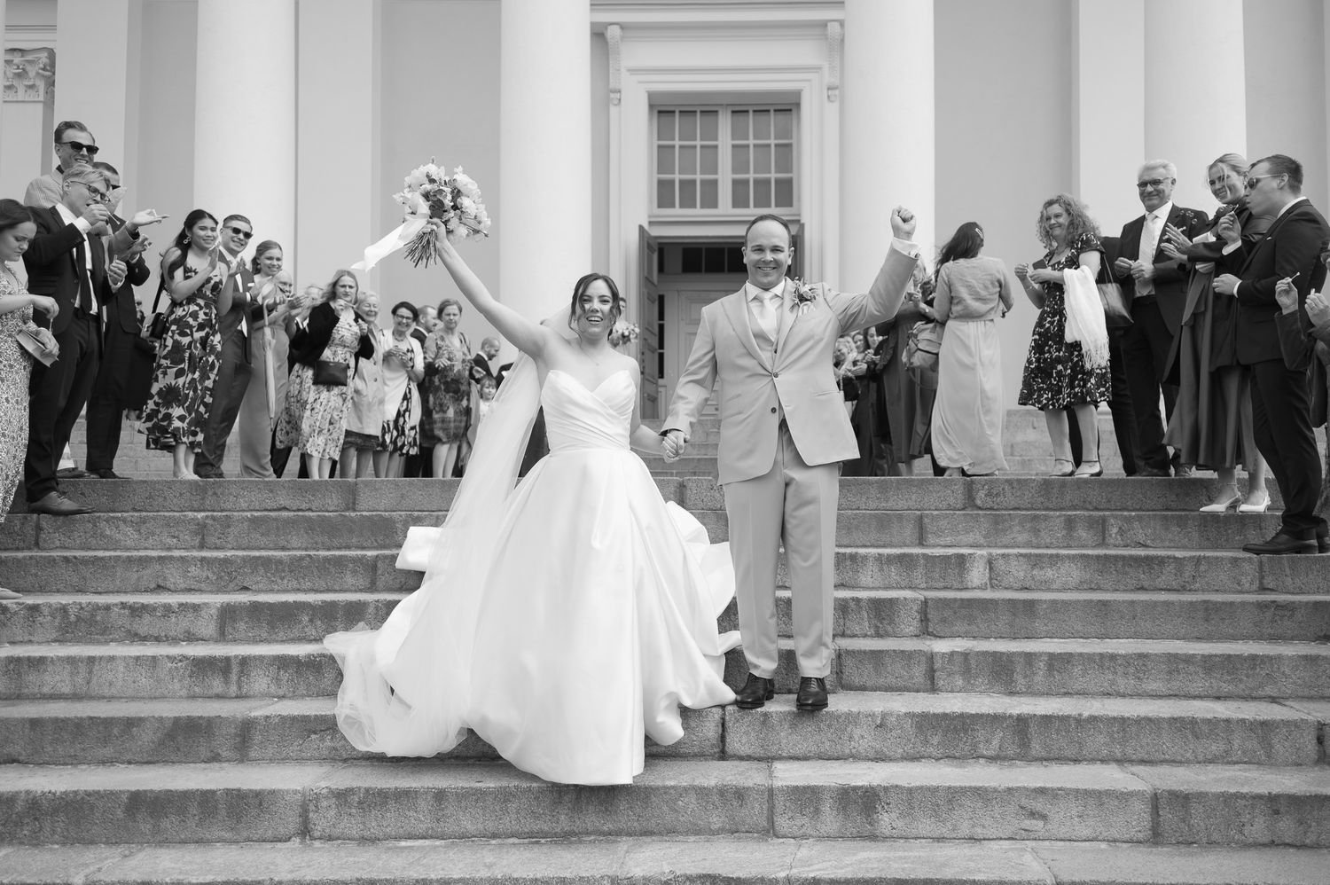 Bride and groom celebrate on grand steps surrounded by cheering guests at elegant outdoor wedding ceremony.