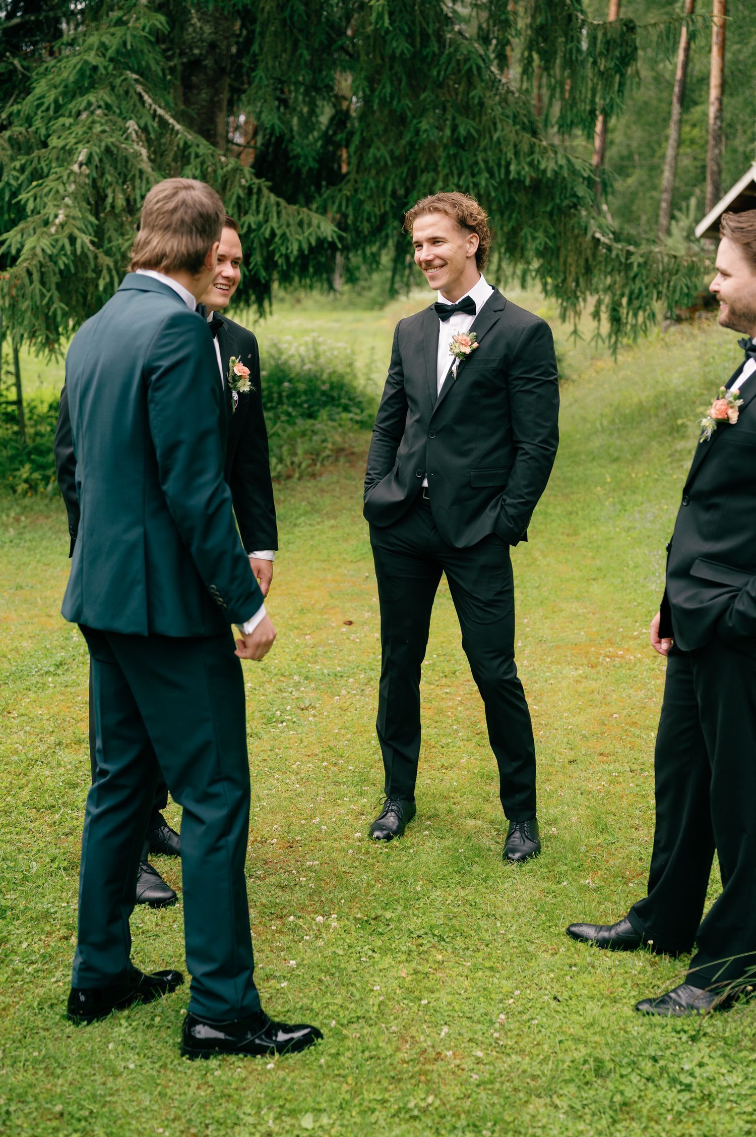 Groomsmen in formal attire stand casually on grass during wedding photos.