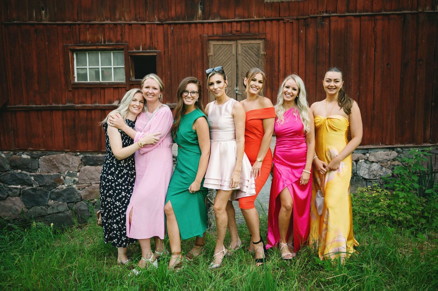 A group in colorful summer dresses poses in front of a red barn wall.