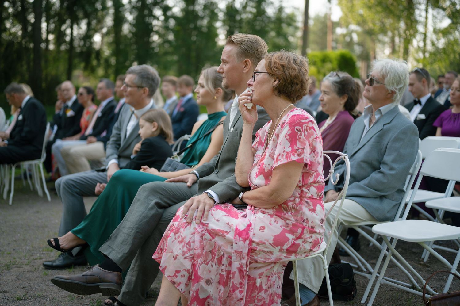 Wedding guests seated in white chairs watching an outdoor ceremony in a garden setting.