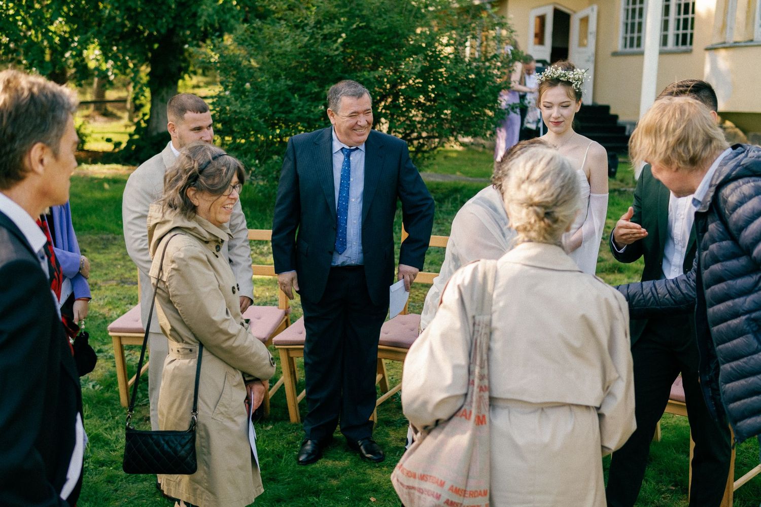 Wedding guests mingle outdoors on a sunny day in a garden setting.