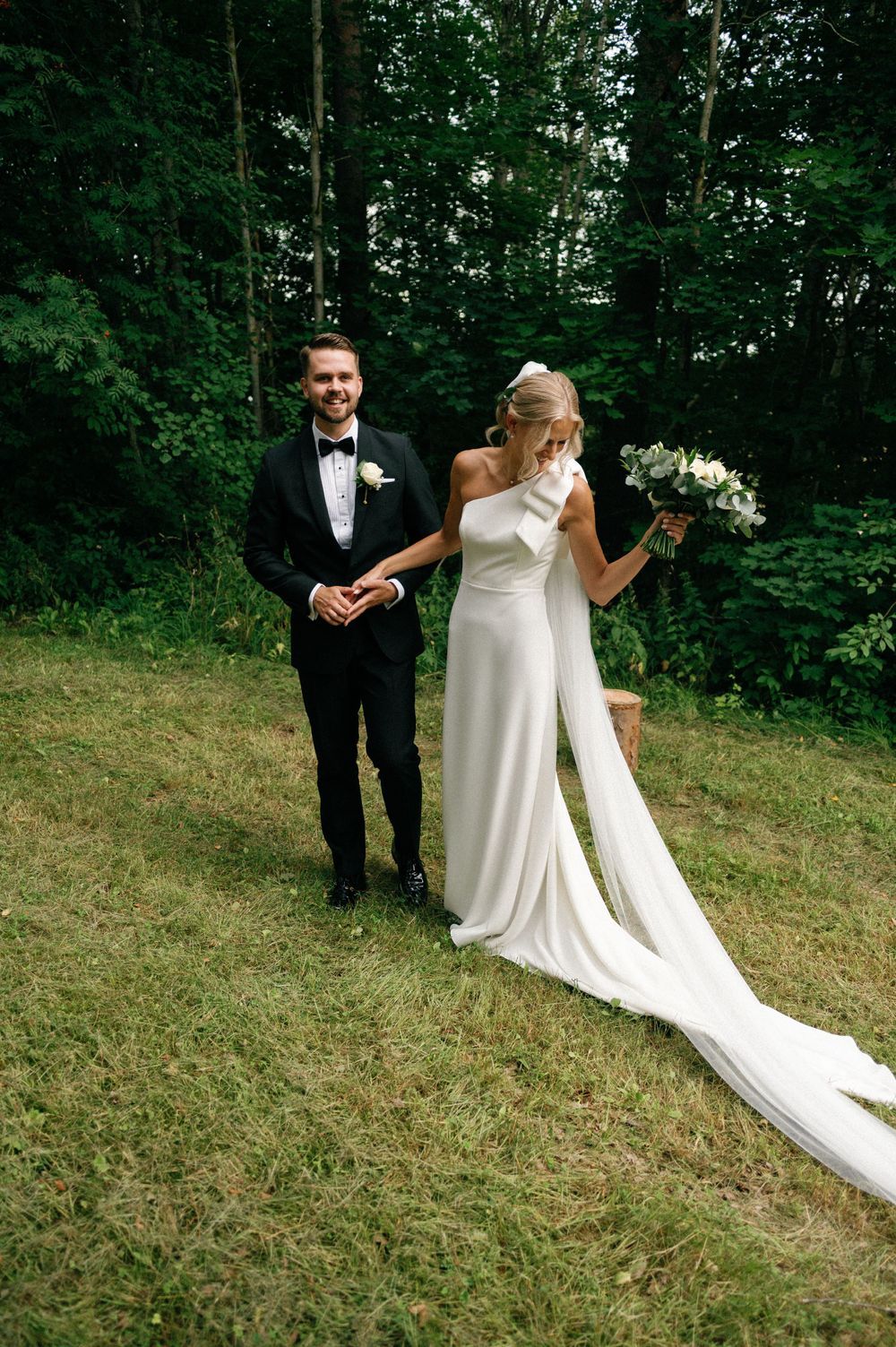 Wedding couple interacts playfully on grassy hillside with long flowing bridal veil.