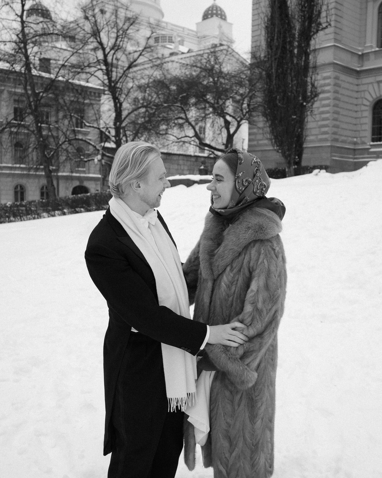 A couple in winter attire share an intimate moment outdoors in the snow with a domed building visible in the background.
