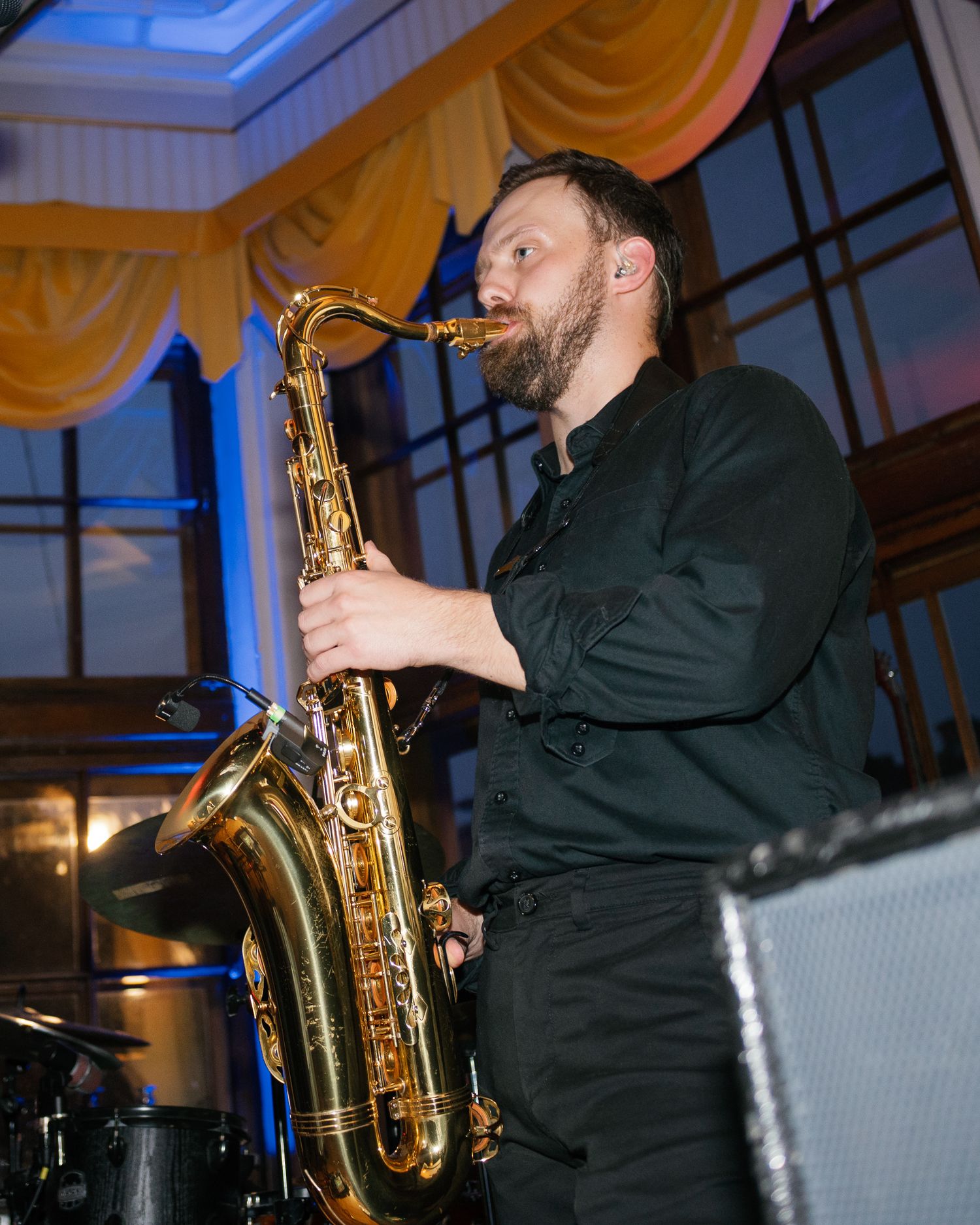 A musician plays a saxophone during a performance in an elegant venue with warm lighting and yellow curtains.