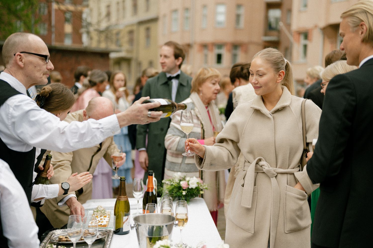 A waiter serves drinks at an outdoor party with guests gathered around a table with wine bottles and glasses.