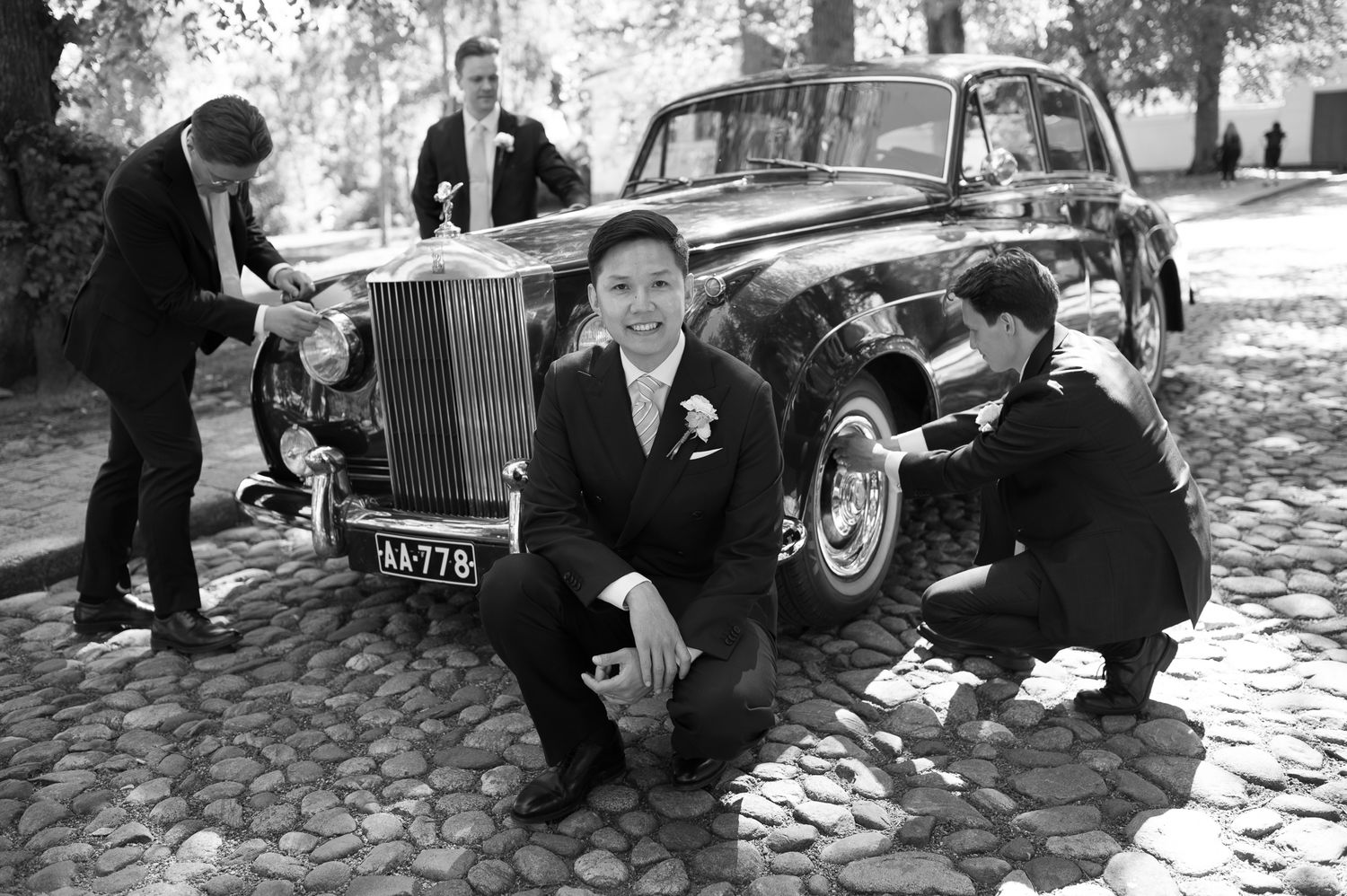 Groom poses in front of vintage Rolls-Royce on cobblestone path while groomsmen polish the classic black wedding car.