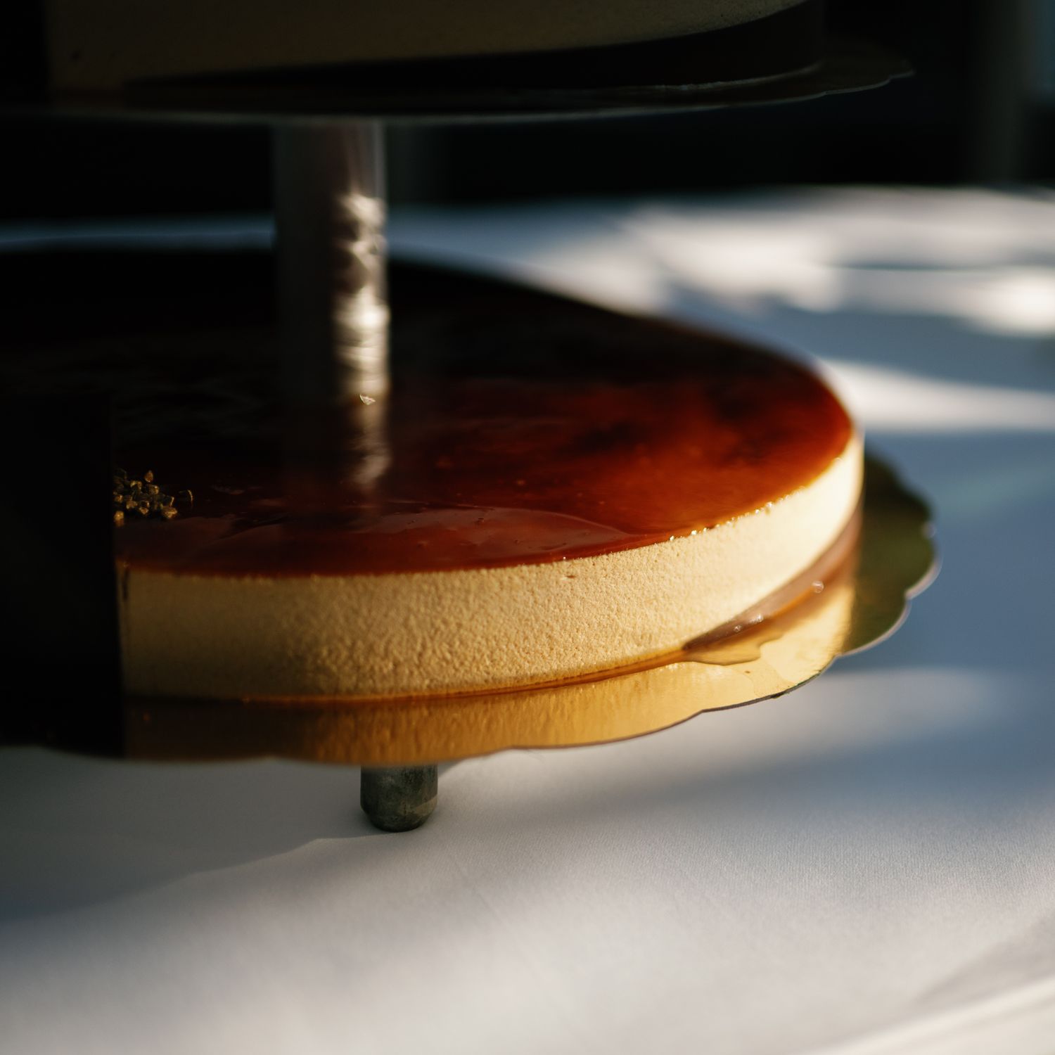 Close-up of a creamy flan dessert with caramel glaze on a golden cake board in dramatic sunlight.