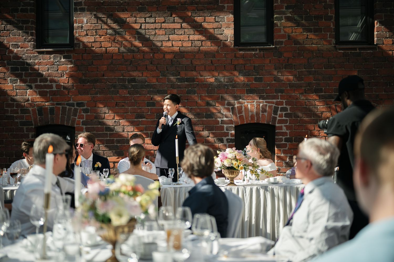 Person giving a speech at an outdoor wedding reception with brick wall backdrop and elegantly decorated tables.