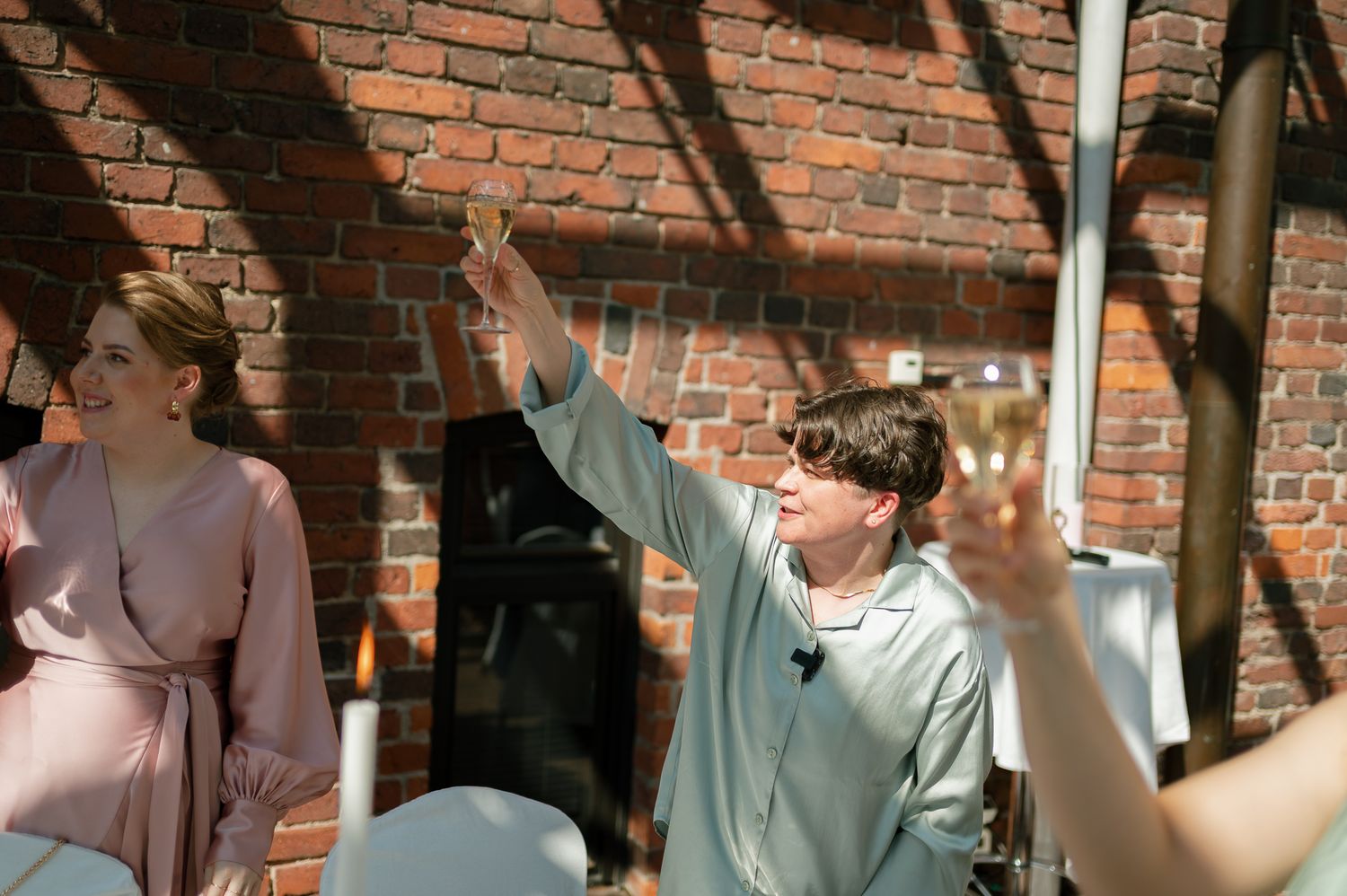 Man raising hand in toast at outdoor wedding reception near brick wall, woman in pink dress smiling beside him.