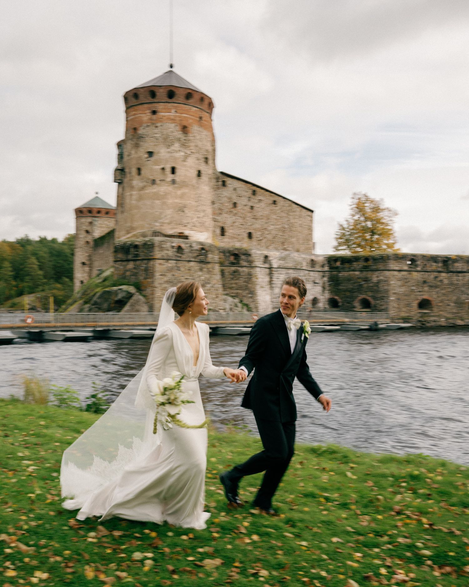 A joyful bride and groom run hand-in-hand on green grass beside a historic stone castle by a river.