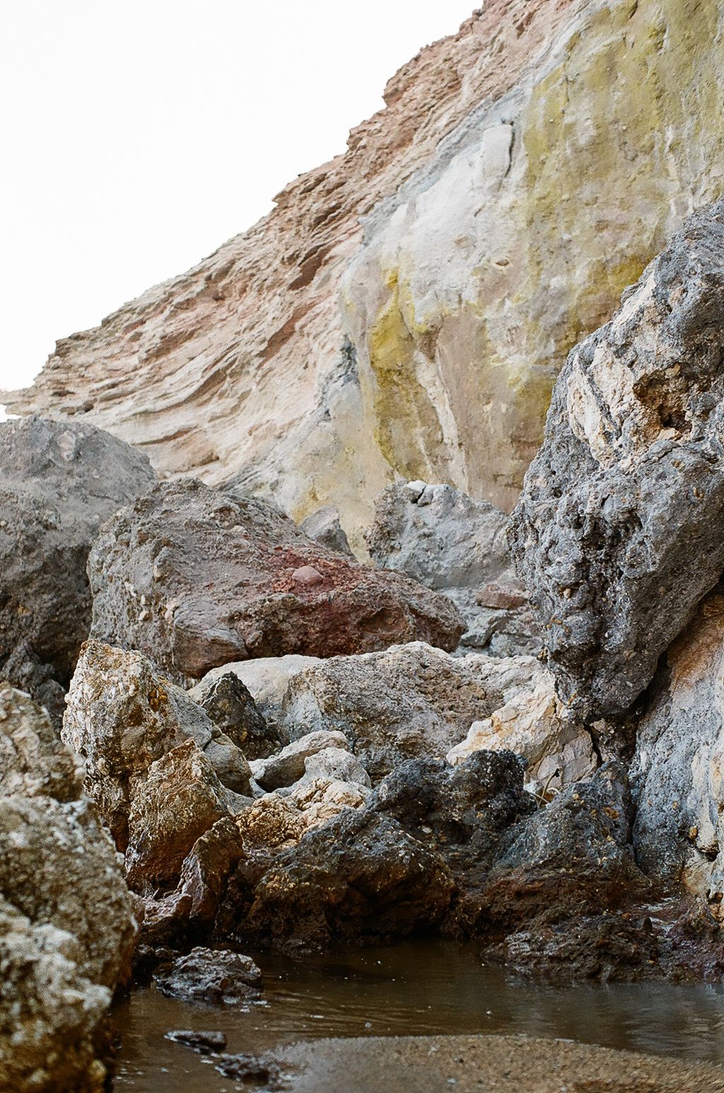 Colorful layered rock cliff with yellow, red, and white mineral deposits above jagged boulders and a small tidal pool.