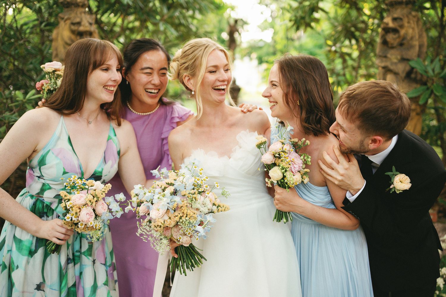 Bride in white gown laughing with bridesmaids in colorful dresses holding floral bouquets outdoors.