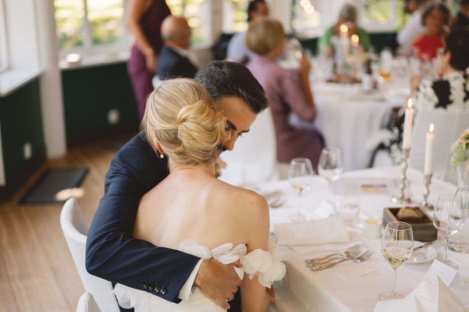 Groom in navy suit embracing bride in white strapless gown at elegant wedding reception dinner table.
