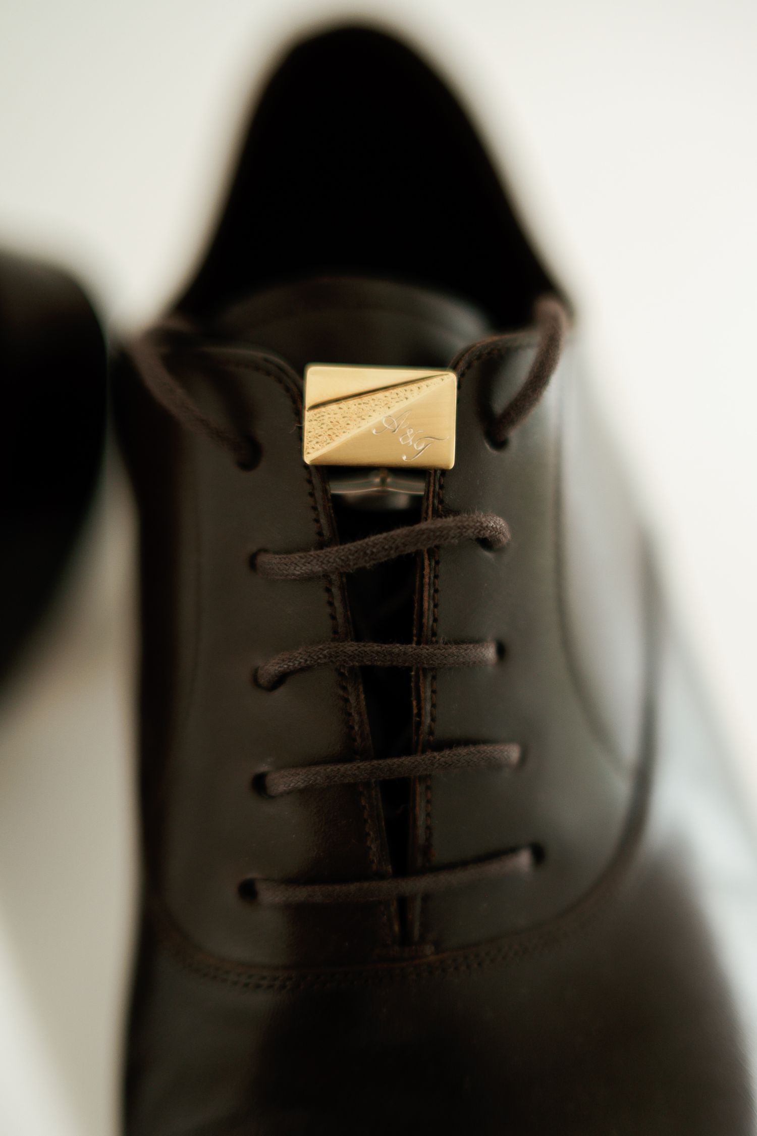 Dark brown leather Oxford dress shoes with brown laces and a gold metal decorative clasp detail on the tongue.