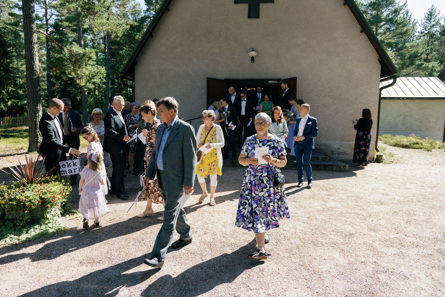 Wedding guests gather outside a rustic chapel with white walls and a cross in a pine forest setting.