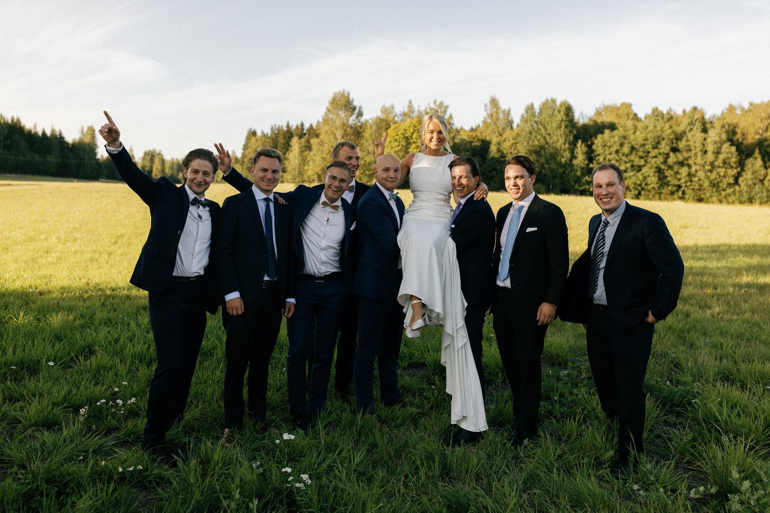 Groomsmen in dark suits celebrate with energetic poses in outdoor wedding photo.