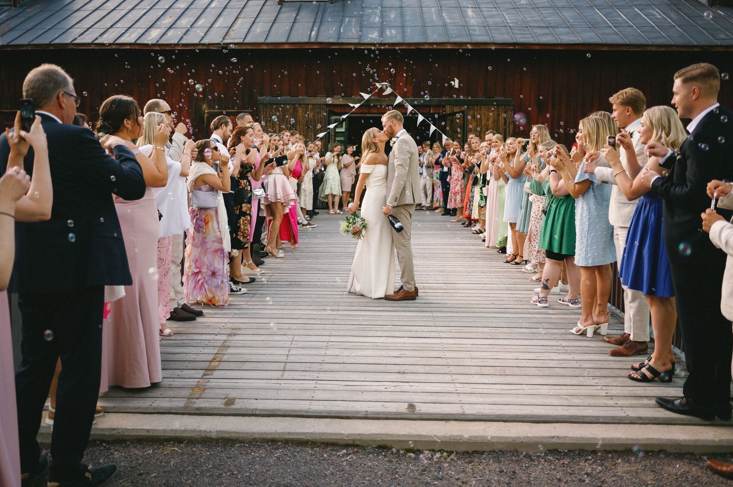 Guests form an aisle on a wooden deck throwing confetti at a wedding celebration.