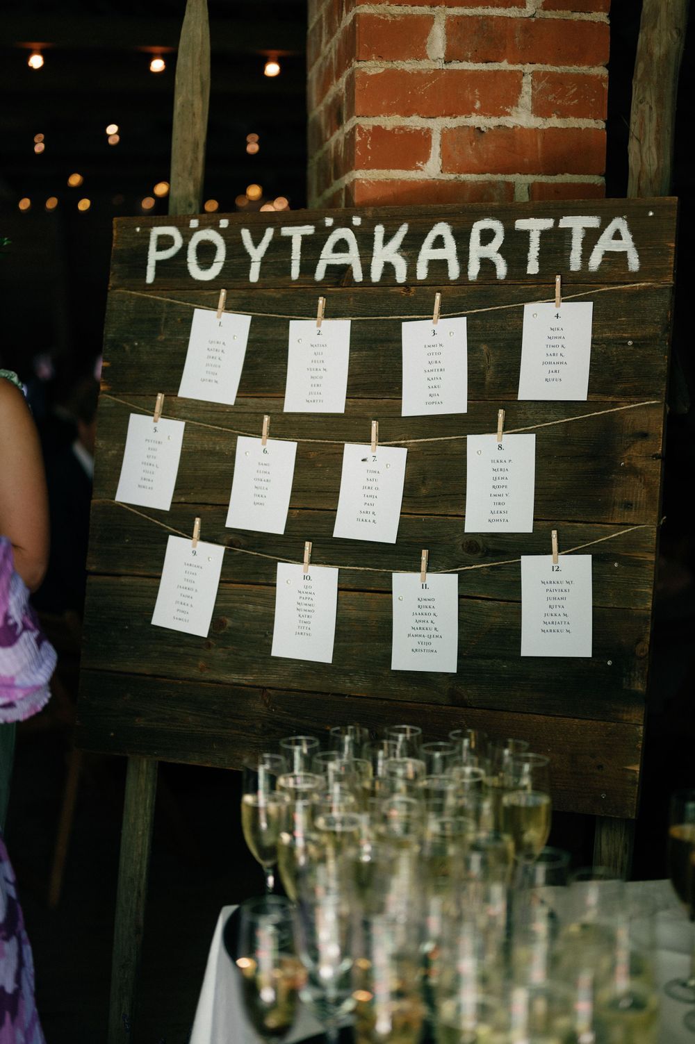 Wedding seating chart display with paper cards hung on strings against a rustic brick wall.