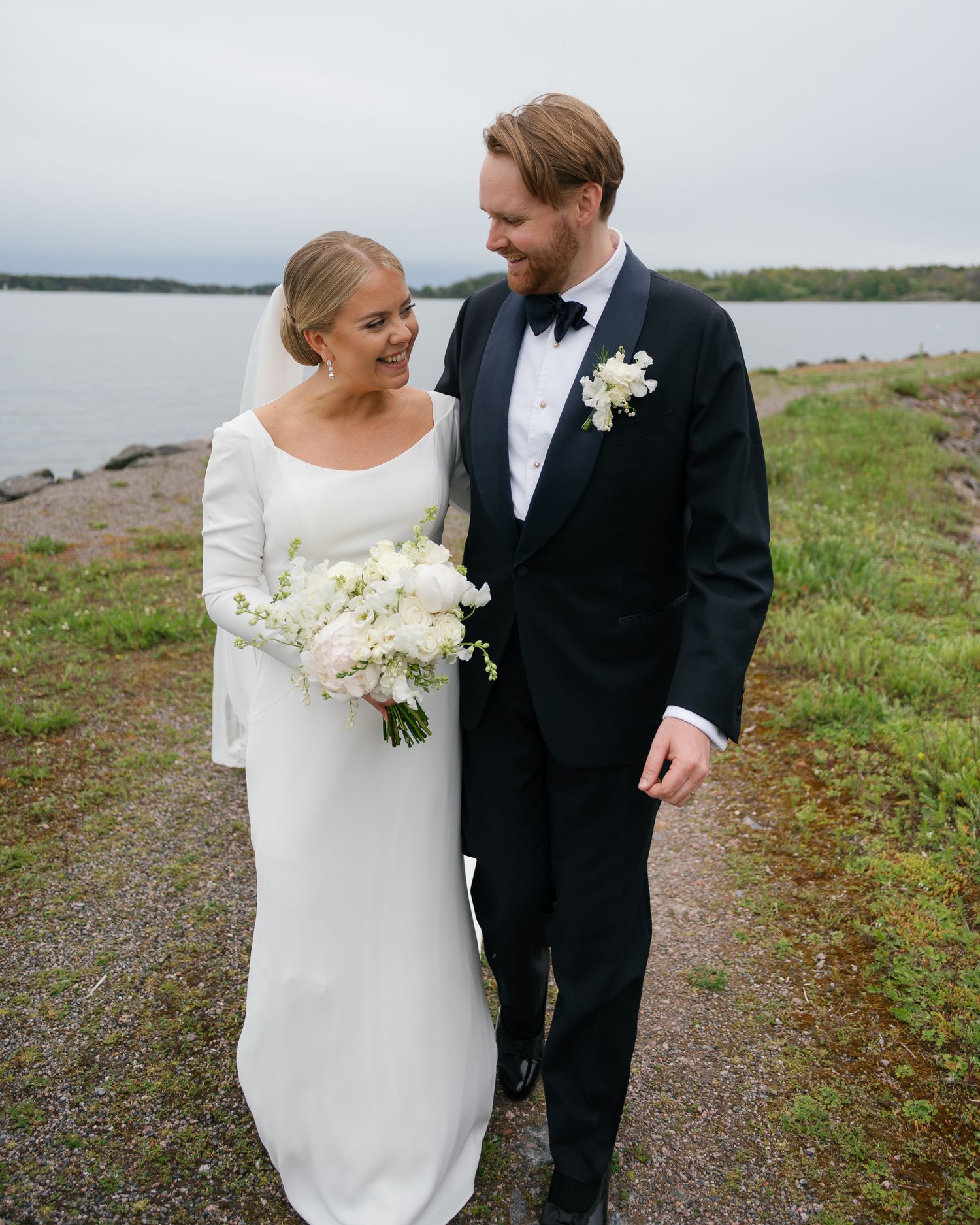 Couple in formal wedding attire walking along a coastal path with water and greenery in the background.