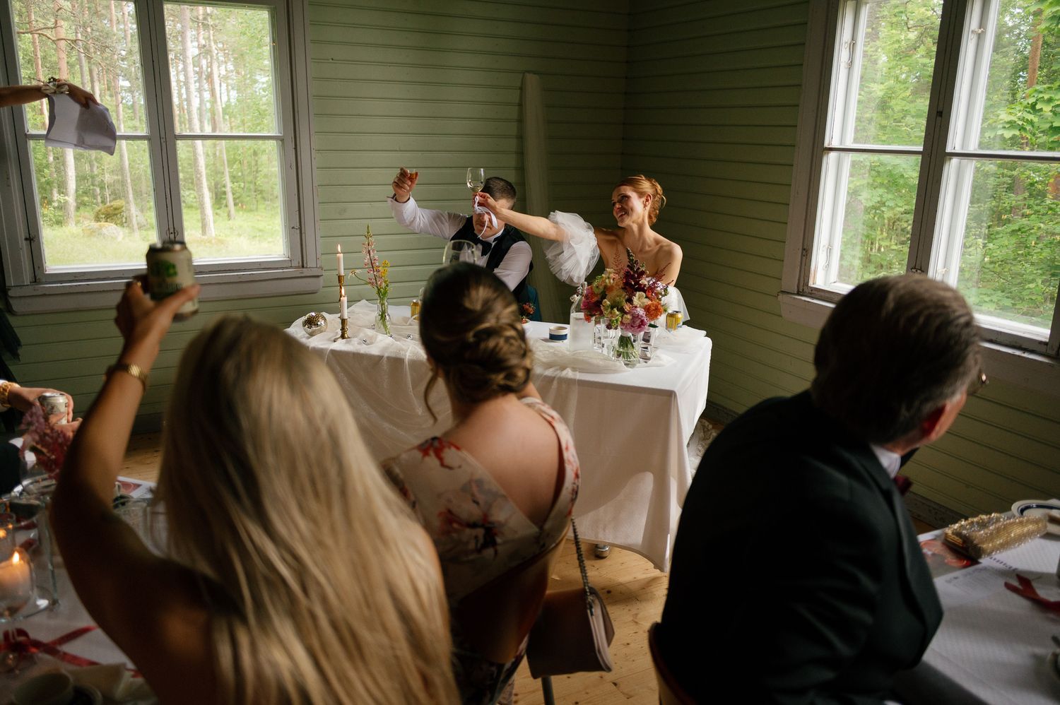 Indoor wedding reception with guests gathered at decorated tables near windows.