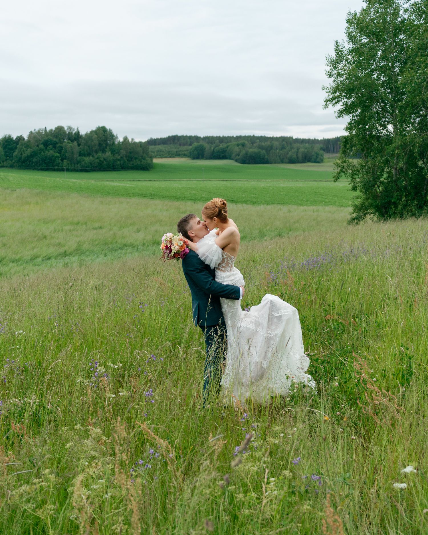 Groom lifting bride in white dress while kissing in a lush green meadow on their wedding day.