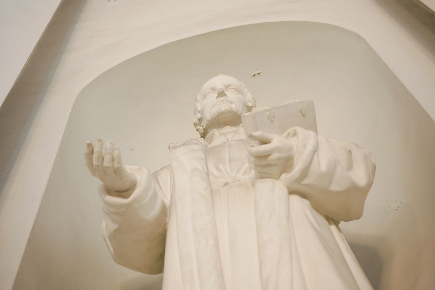White marble statue of a robed religious figure holding a book with outstretched arm in an arched niche.