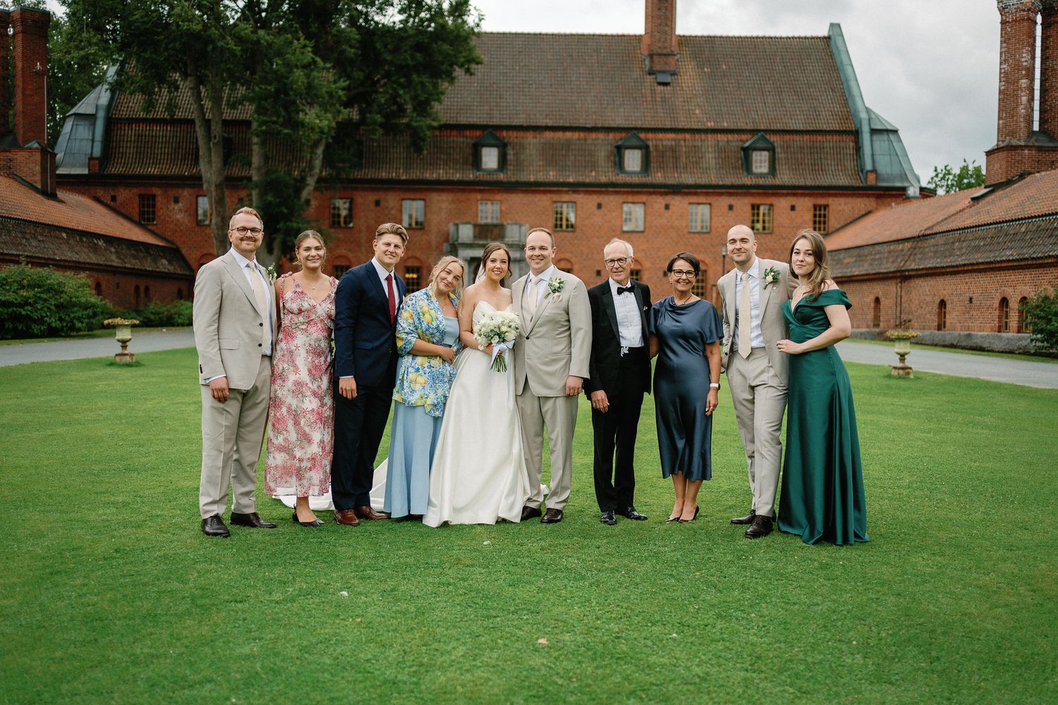 Wedding party poses on green lawn before red brick venue, bride in white gown flanked by family in formal attire.