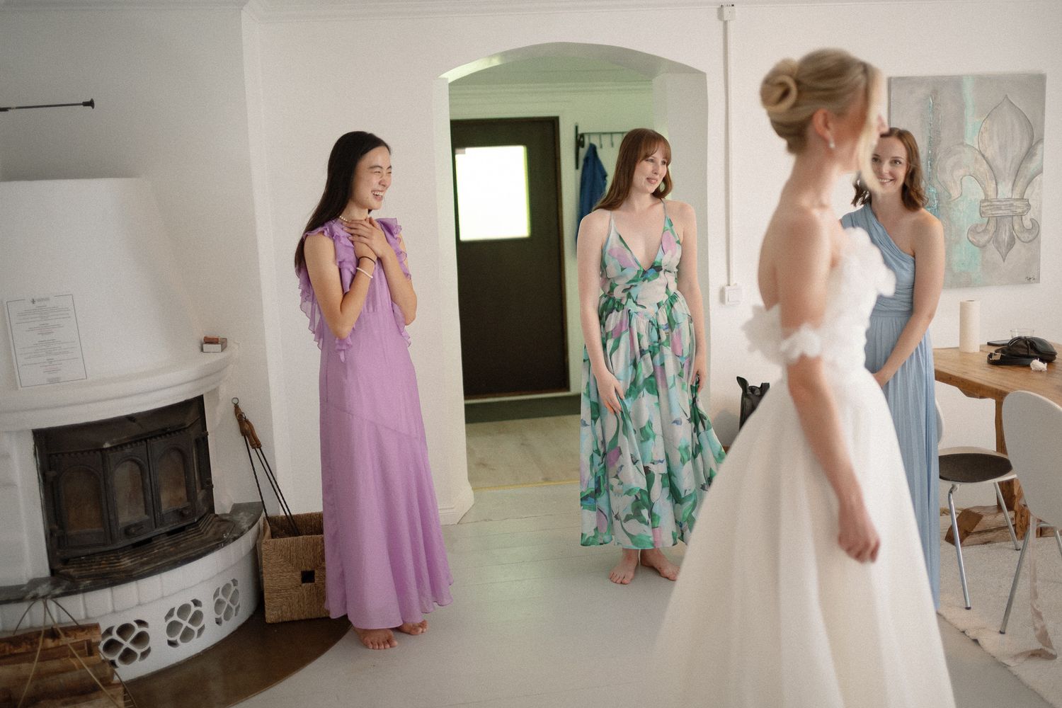 Bride in white wedding gown shows dress to three bridesmaids in colorful dresses inside a bright white room.