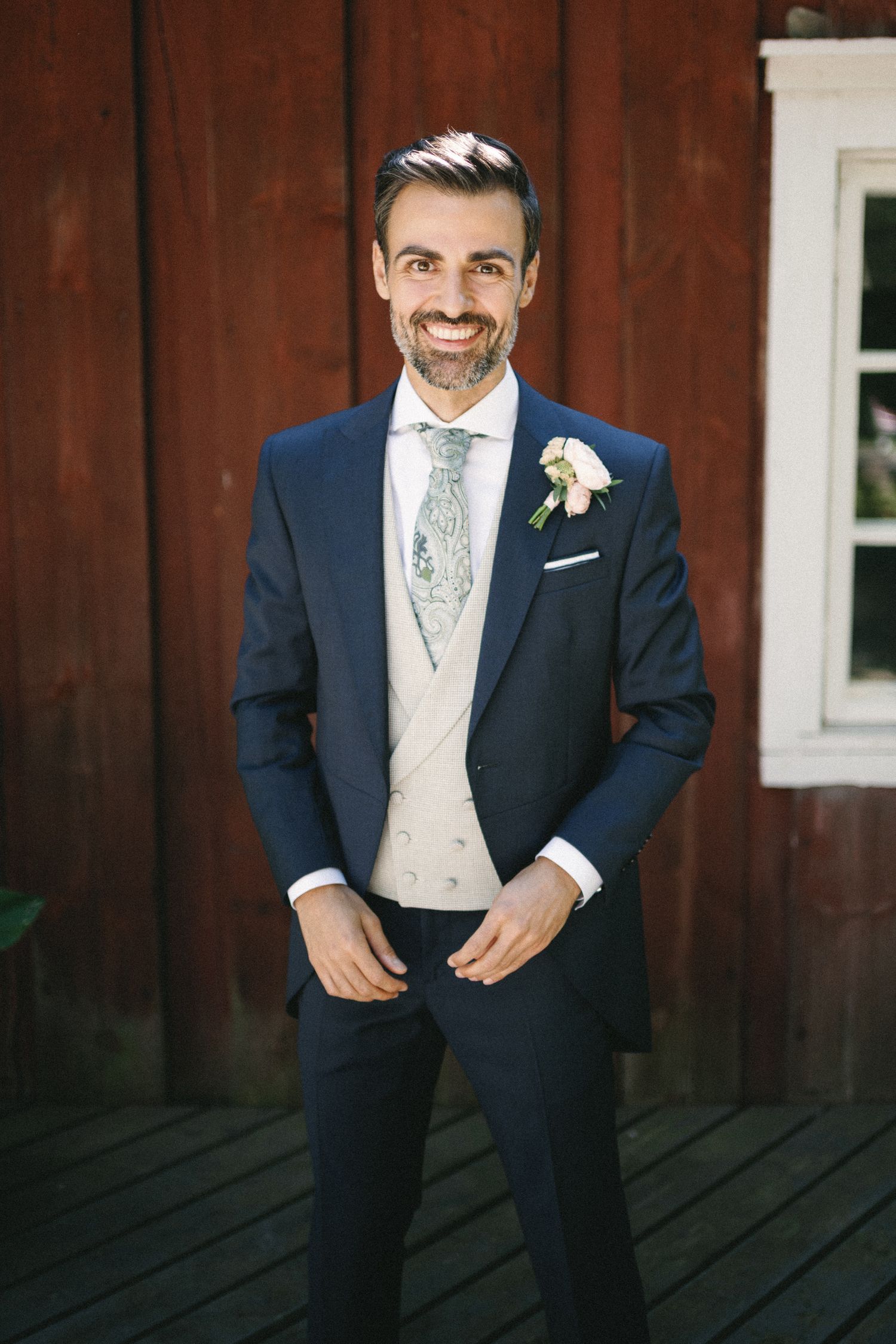 Smiling groom in navy suit with boutonniere and patterned tie poses outdoors near red barn.