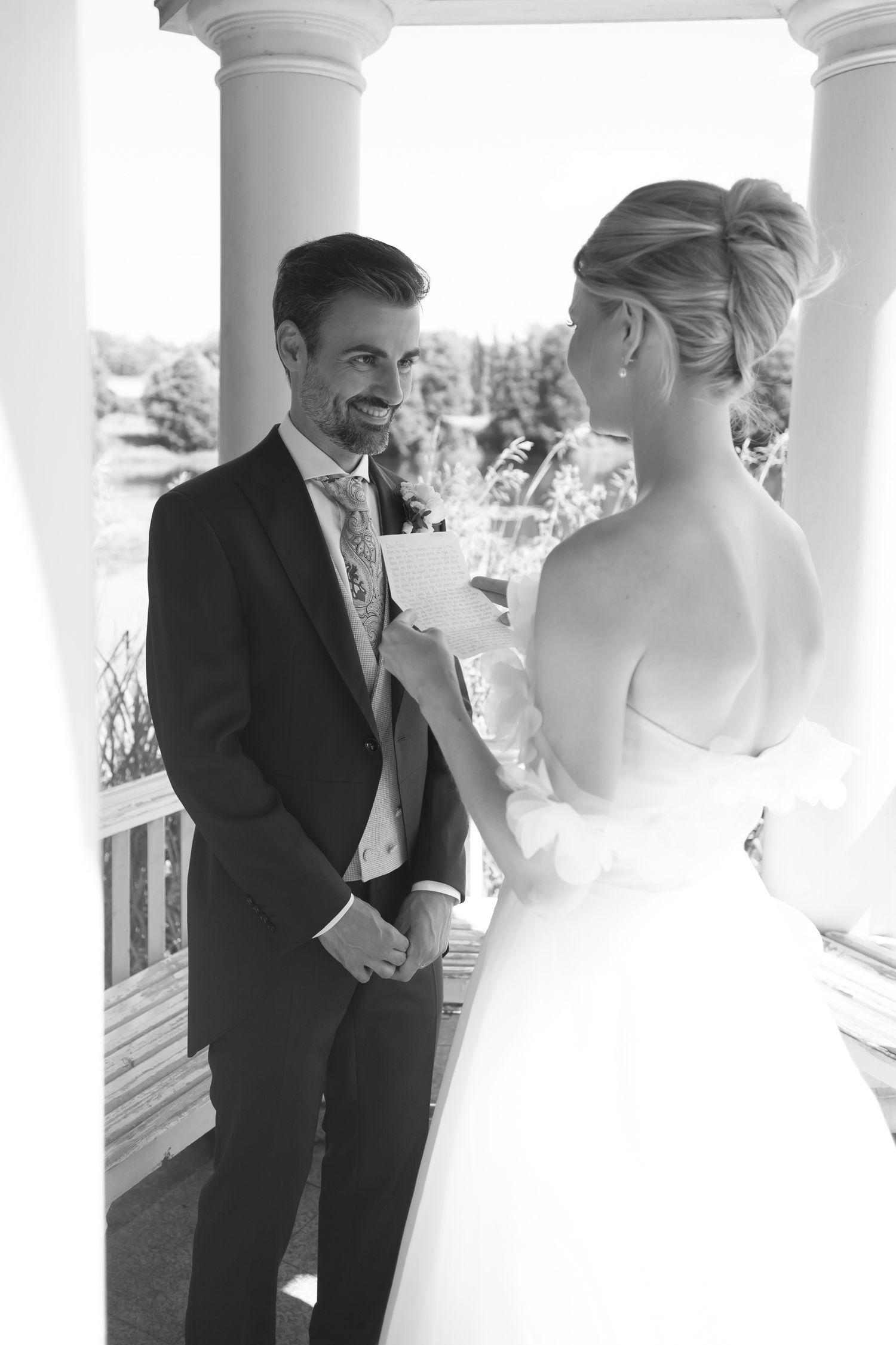 Black and white photo of a bride reading wedding vows to a smiling groom on a columned porch.