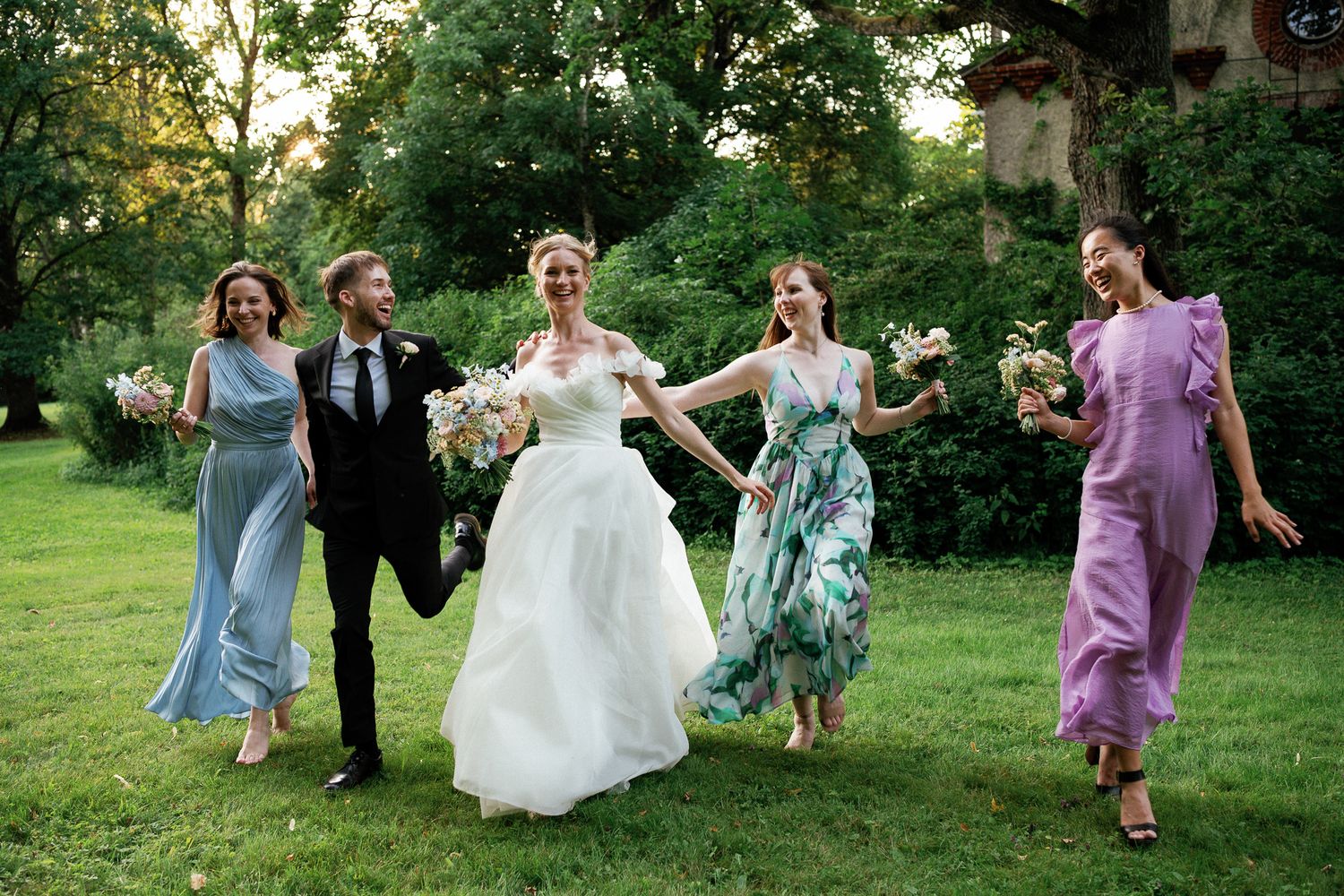 Bride and groom walk with three bridesmaids in colorful dresses on lush green lawn during outdoor wedding celebration.
