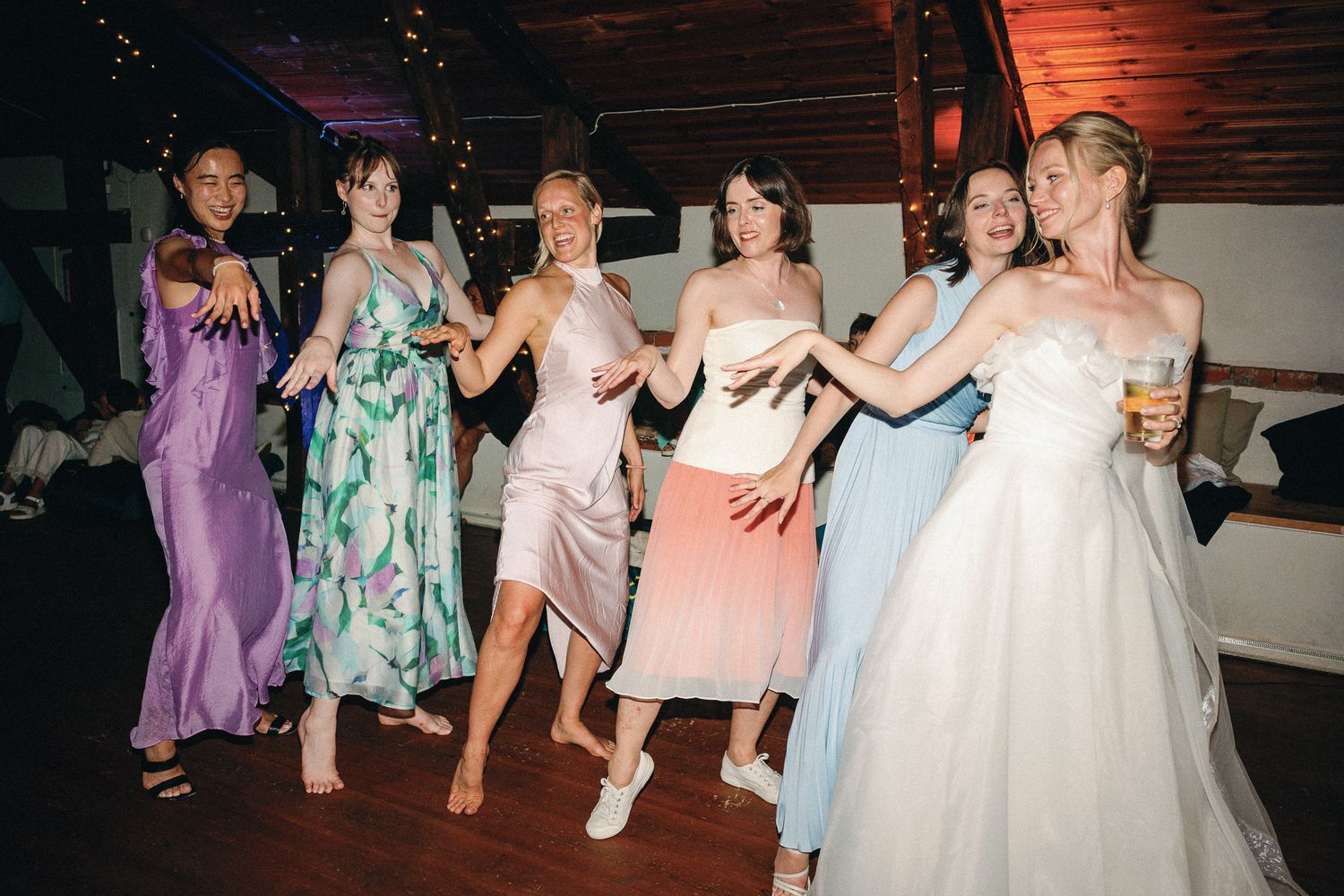 Six women in elegant dresses dancing joyfully at a wedding reception on a wooden dance floor.