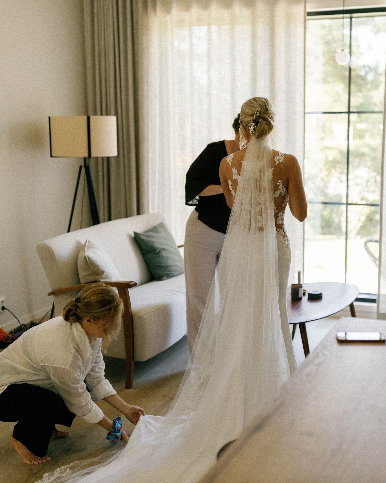 A wedding dress being carefully arranged on a bed in a bright sunlit room.